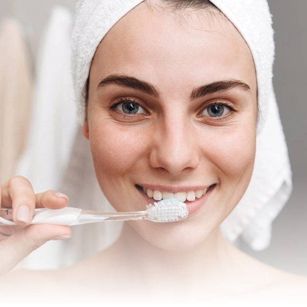 Mulher jovem com toalha na cabeça escovando os dentes, exemplificando cuidados pessoais com a saúde bucal.