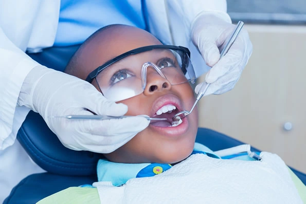 Criança em consulta odontológica para limpeza e prevenção de cáries, usando óculos de proteção enquanto o dentista realiza a profilaxia para remoção de placas e controle de manchas iniciais.